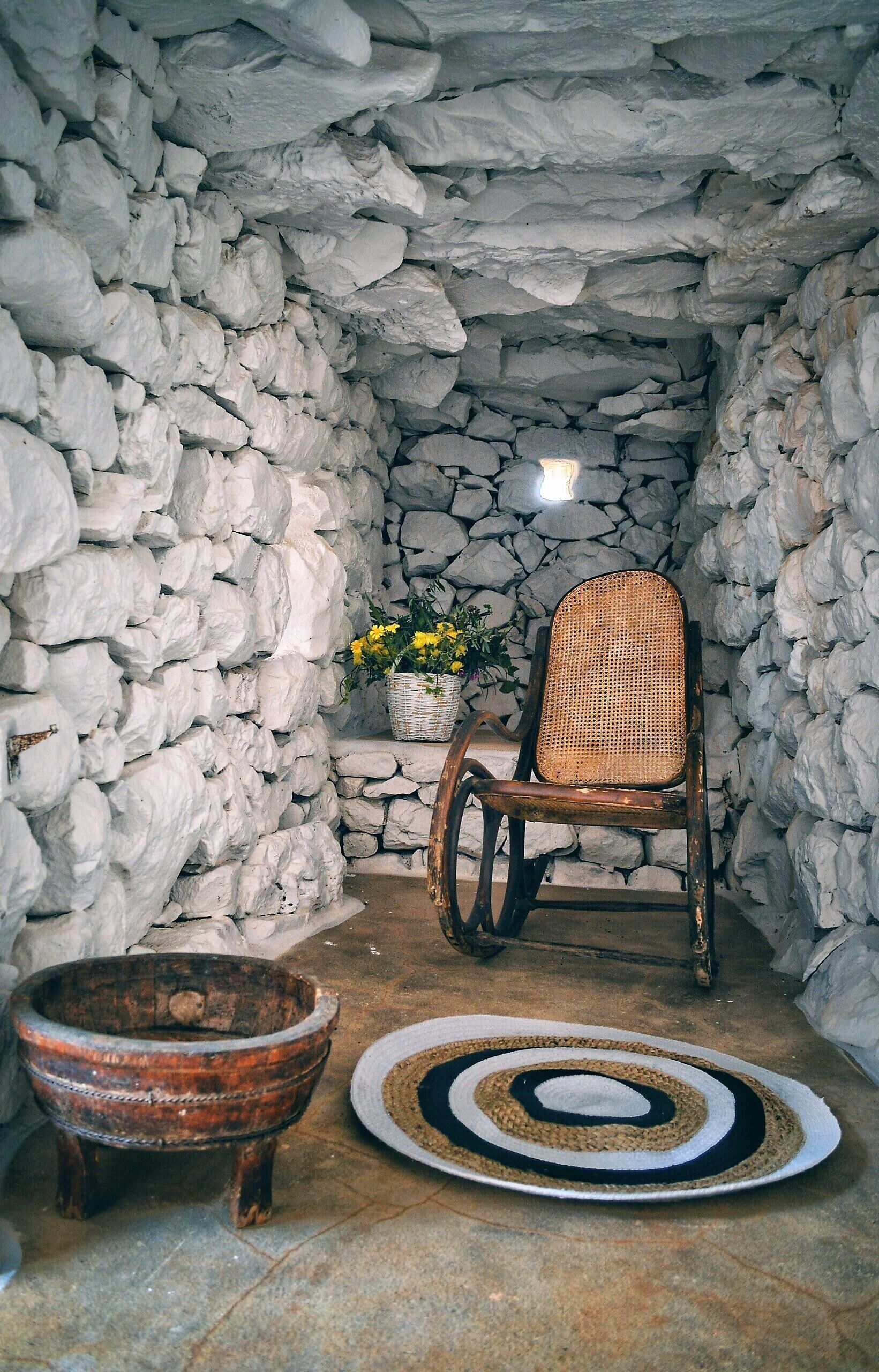 A stone-walled room with a rustic wooden chair, a wicker basket with flowers, and a woven rug.