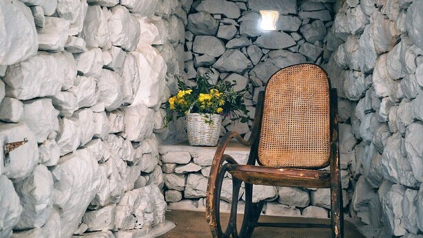A stone-walled room with a rustic wooden chair, a wicker basket with flowers, and a woven rug. - Lithos Stone Suites (East Mani)