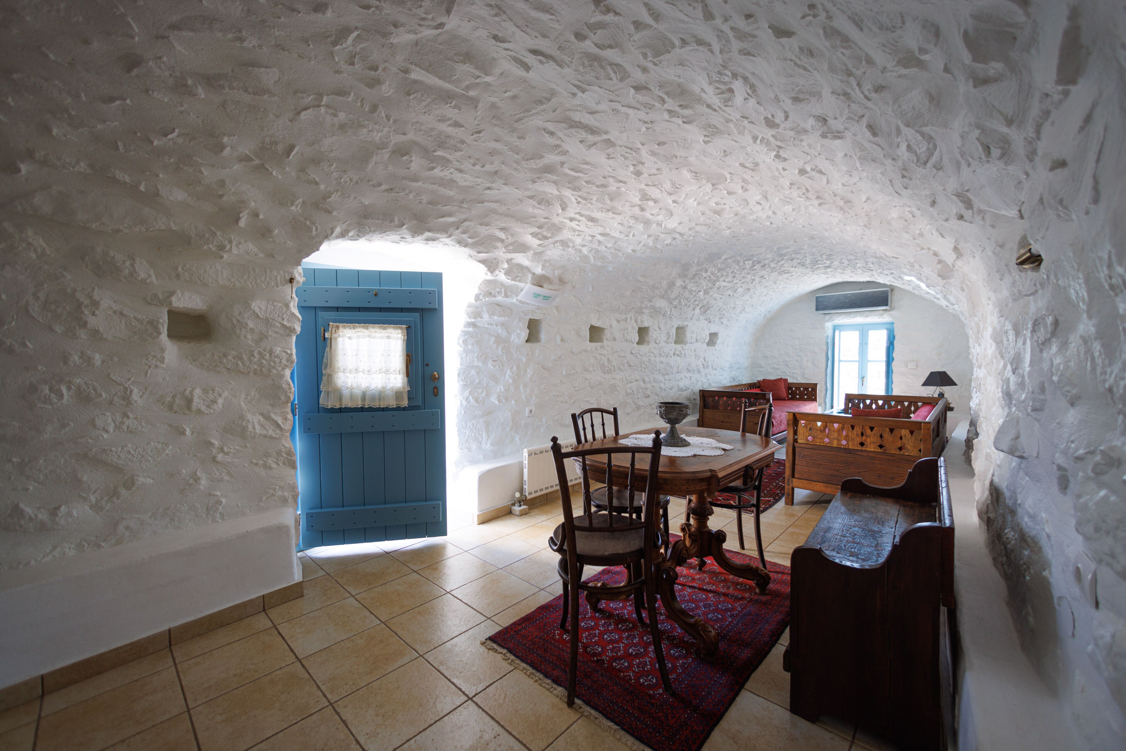 A rustic interior with a stone wall, a blue door, a wooden table with chairs, and a patterned rug.