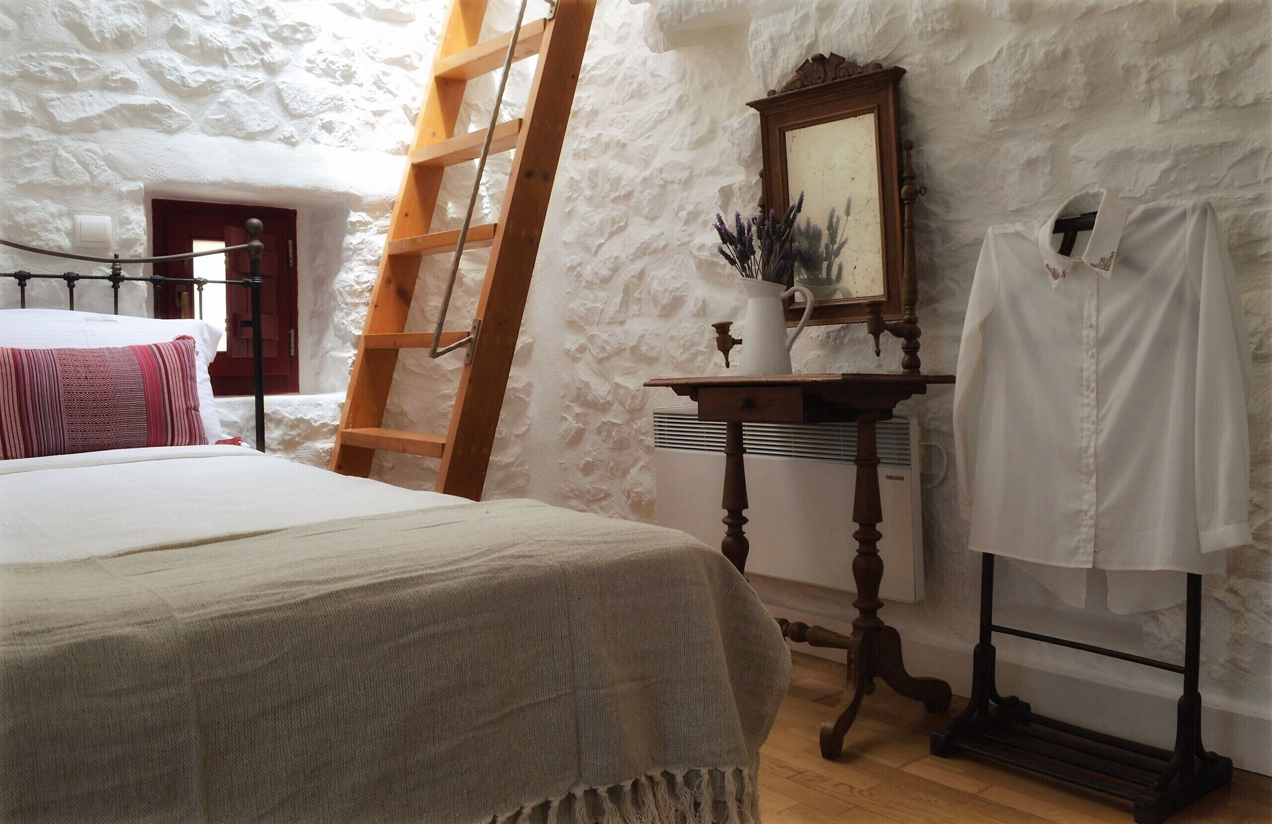 A bedroom with a stone wall, a wooden ladder, a bed with a white and red blanket, a small table with a vase, and a white shirt on a hanger.