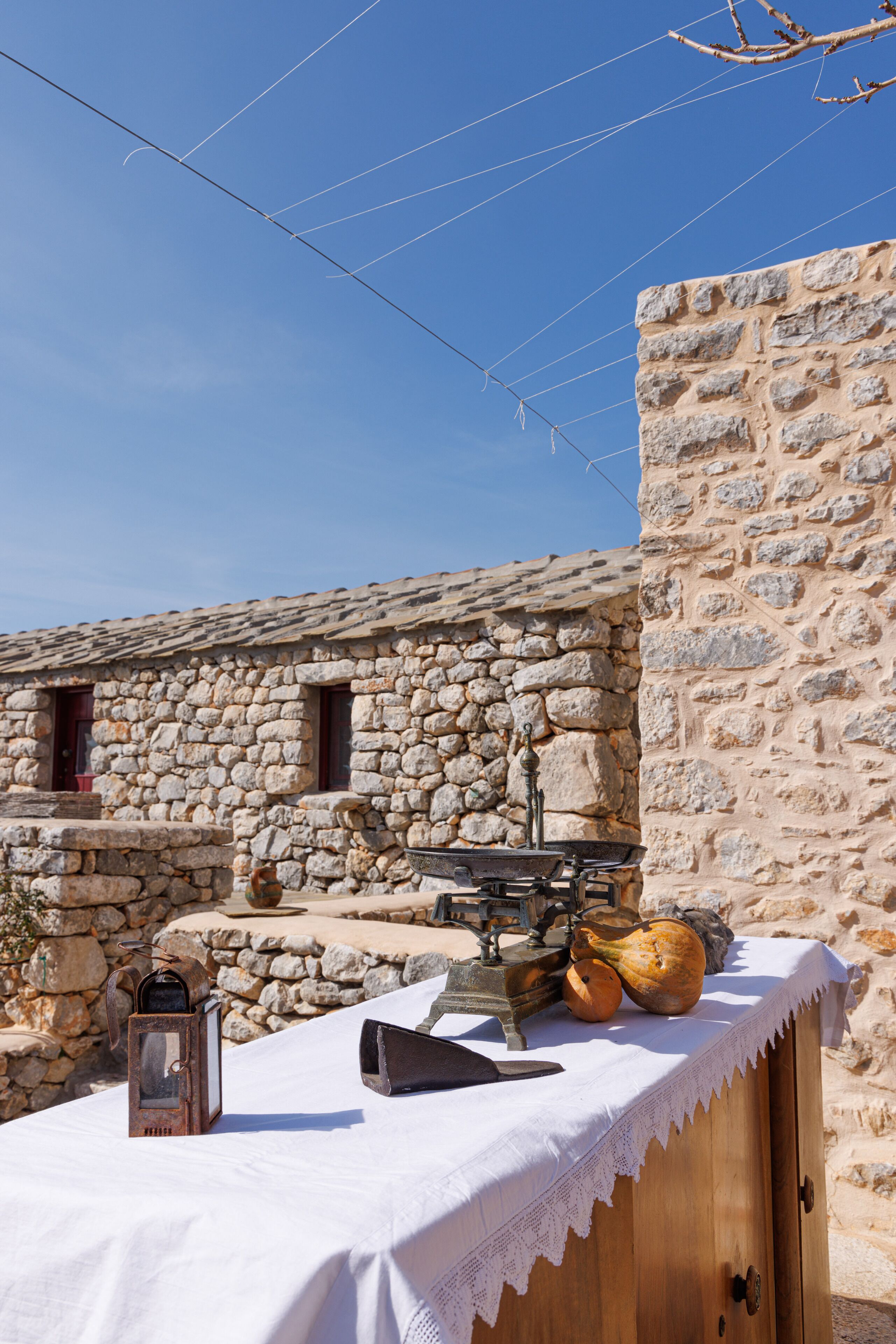 A rustic stone building with a wooden table covered by a white cloth, featuring a lantern, a knife, and pumpkins.