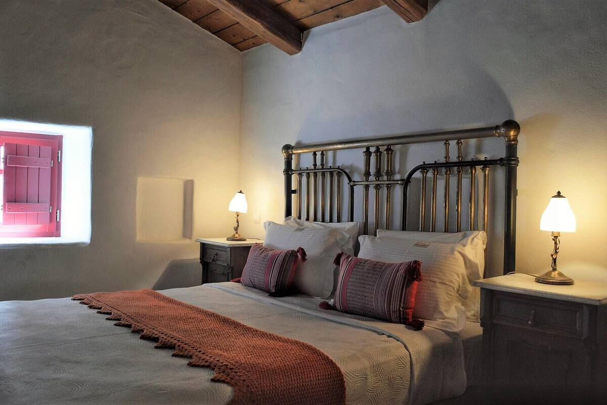 A bedroom with a bed, two bedside tables with lamps, a window with a view, and a wooden ceiling.