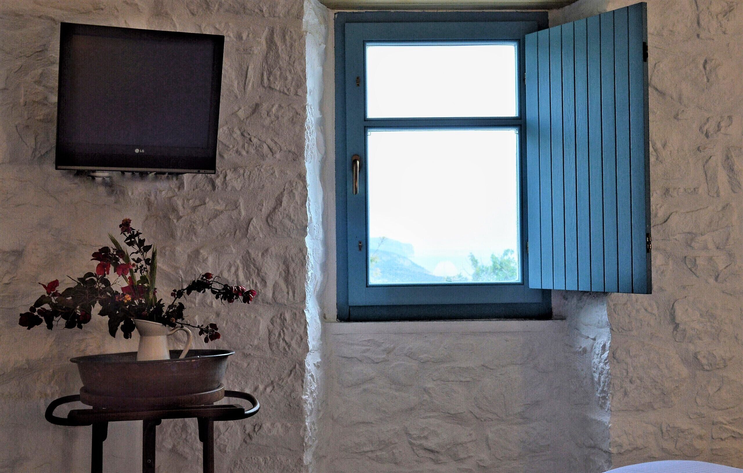 Una habitación con pared de piedra, televisor sobre una mesa con un florero y ventana con contraventanas azules.