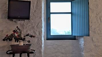 Una habitación con pared de piedra, televisor sobre una mesa con un florero y ventana con contraventanas azules.
