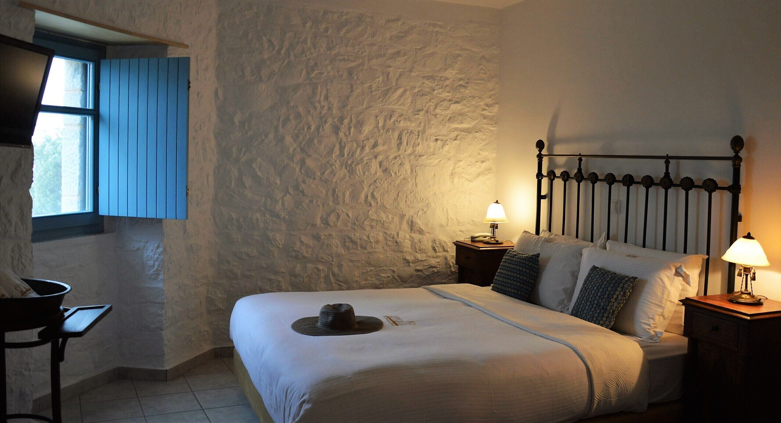Een slaapkamer met een stenen muur, een bed met kussens, een nachtkastje met een lamp en een raam met blauwe luiken.