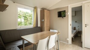 Een compacte kamer met een houten eettafel, witte stoelen, een grijze bank, een aan de muur bevestigde televisie, en een deur naar een slaapkamer.