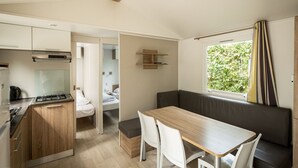Een kleine keuken met een fornuis, een zithoek met een tafel en stoelen, een bankje en een raam met gordijnen.