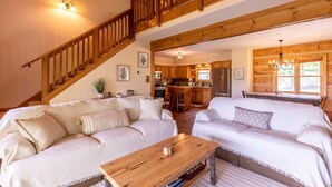 Amplia sala de estar con una escalera de madera, dos sofás, una mesa de centro de madera y una cocina al fondo.