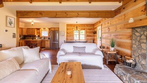 Interior de una cabaña de troncos con sala de estar, cocina y comedor, que presenta vigas de madera, chimenea de piedra y un ambiente acogedor.