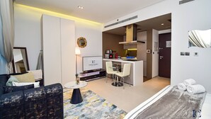 A modern hotel room with a sofa, a small round table, a TV, a kitchen area with a bar, and a bed with towels. - AST - Modern studio flat with balcony (Dubai)