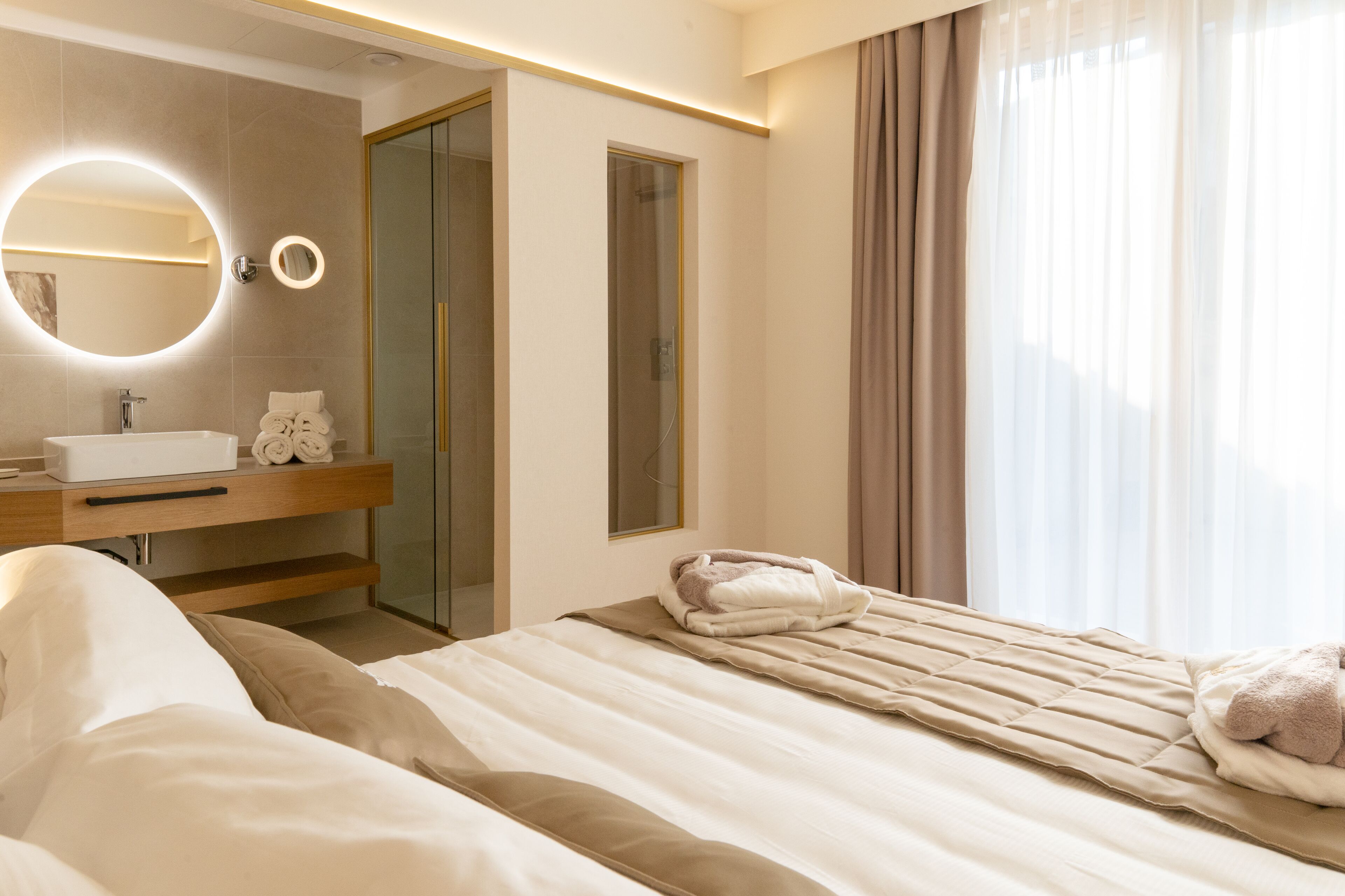 A modern hotel room with a bed, a bathroom with a sink and mirror, and a glass shower door.