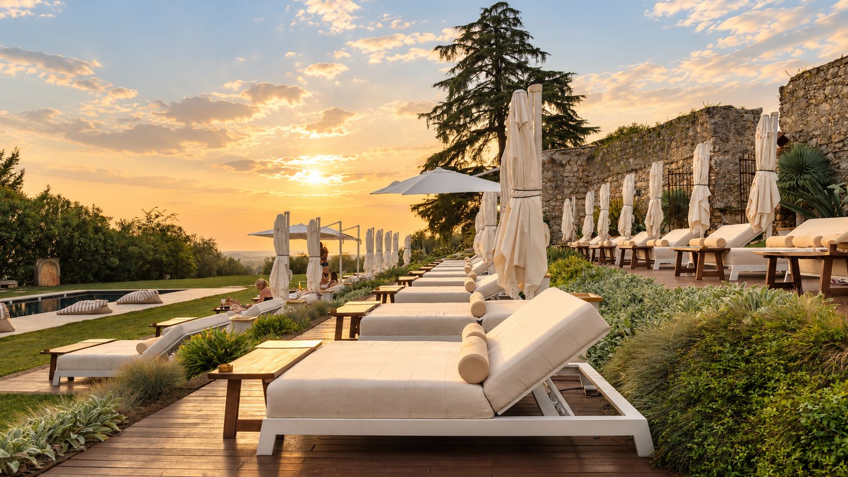 A sunlit outdoor lounge area with sunbeds and umbrellas, a stone wall, and a pool in the background.