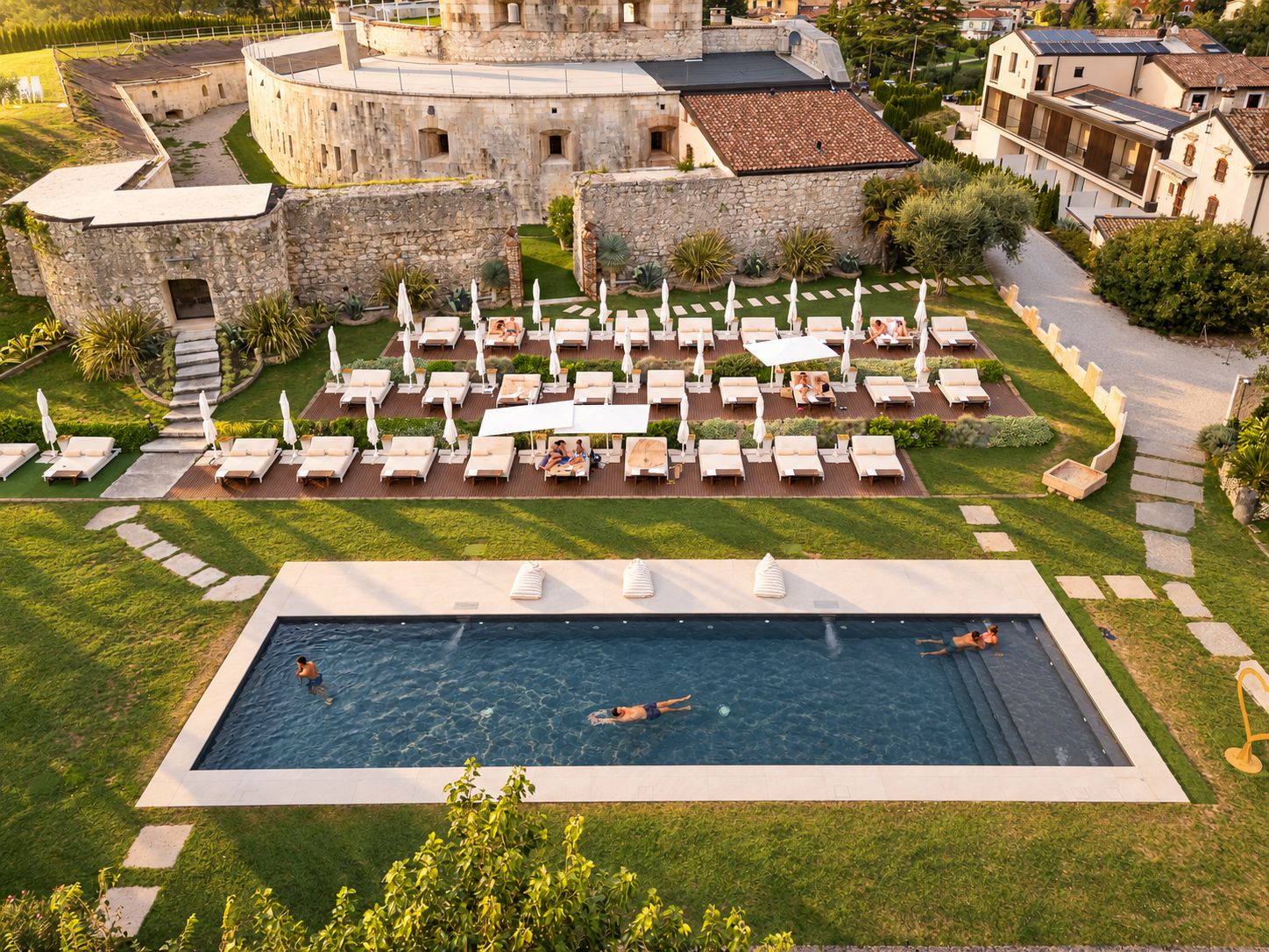 Un espace piscine agrémenté de chaises longues et de parasols, entouré d’un bâtiment en pierre et de verdure.