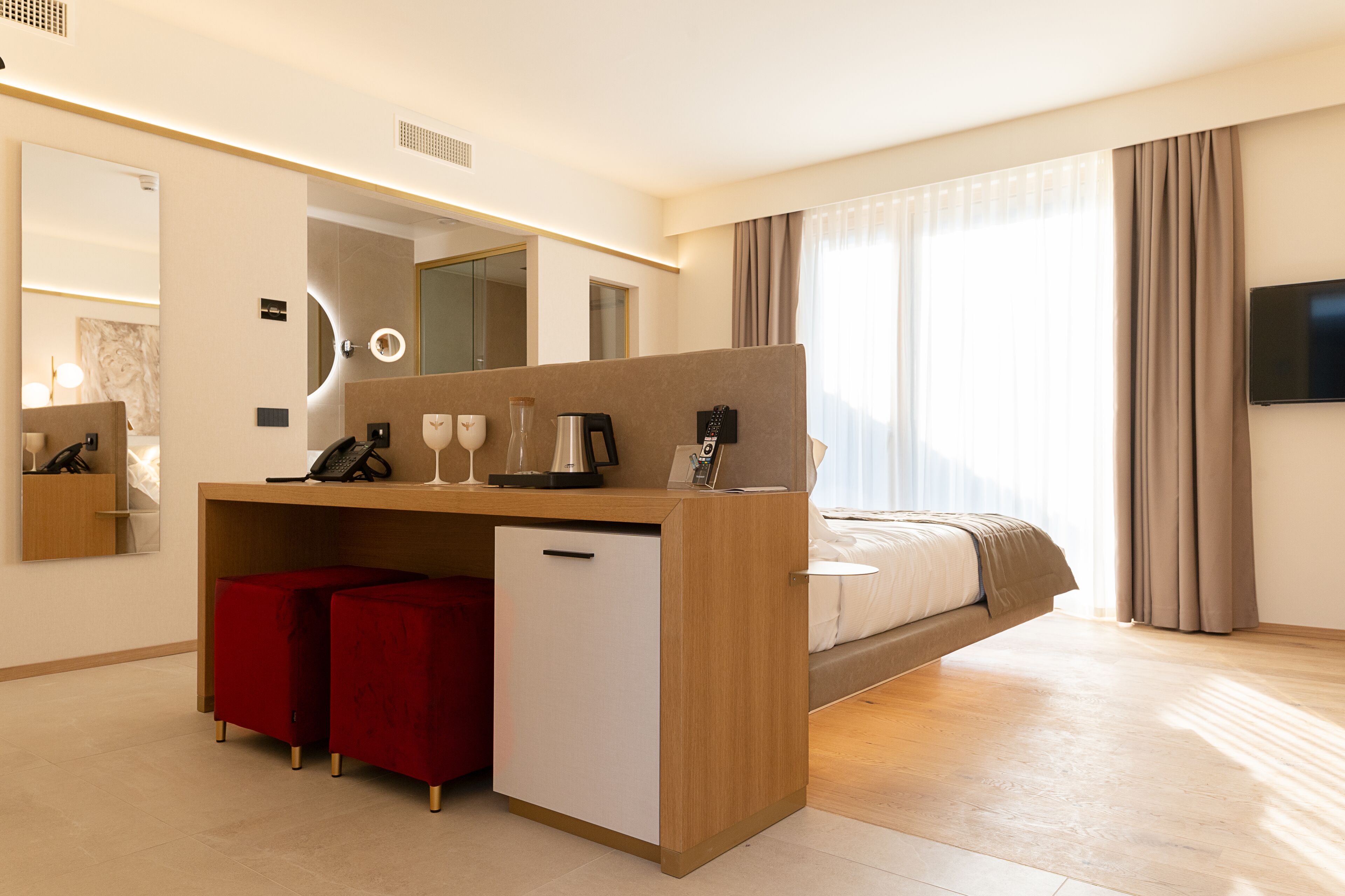 Una habitación de hotel moderna con una cama, un escritorio con teléfono y cafetera, un refrigerador pequeño y un espejo grande.