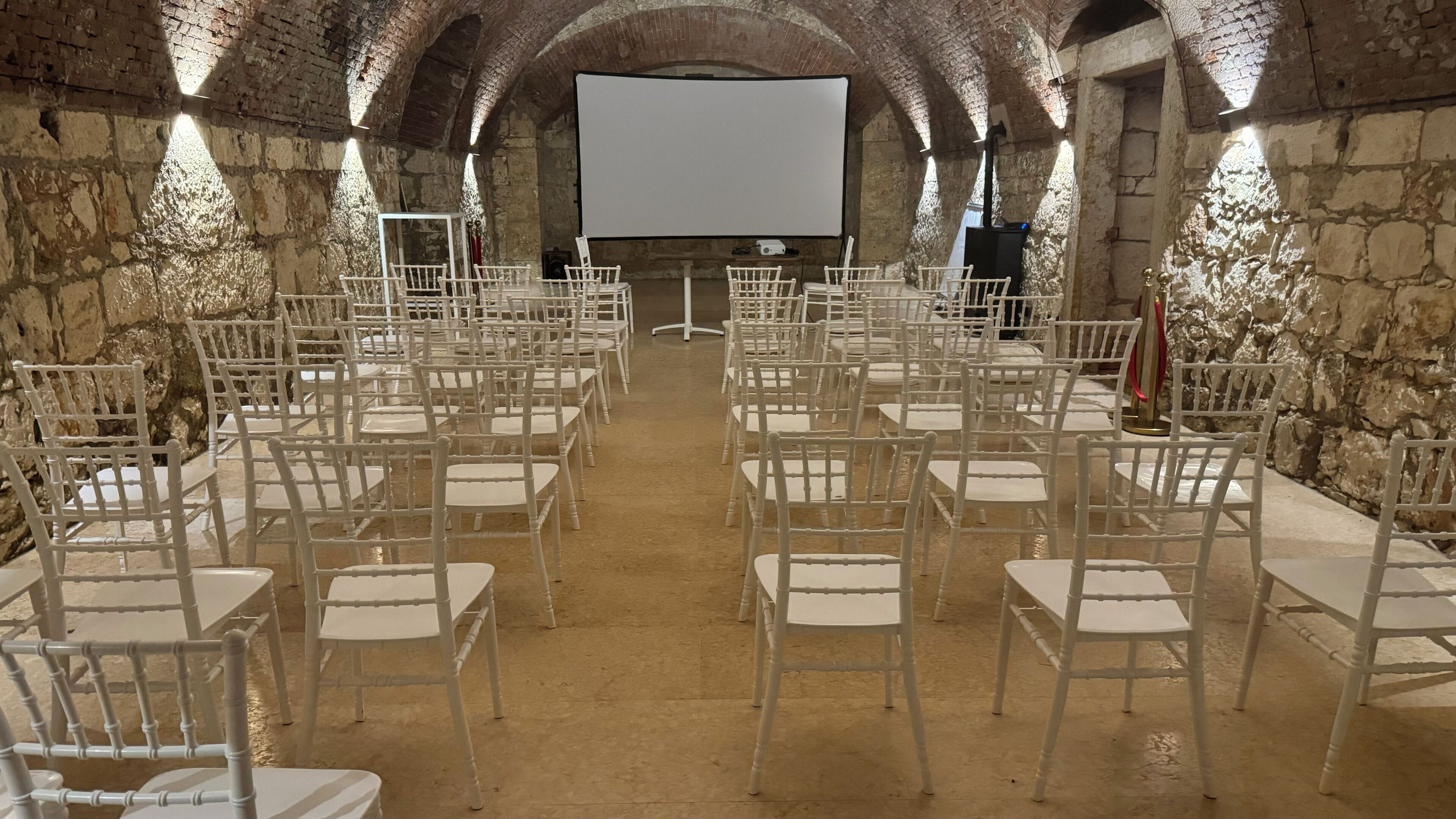 An indoor venue with rows of white chairs and tables, a large screen at the front, and stone walls.