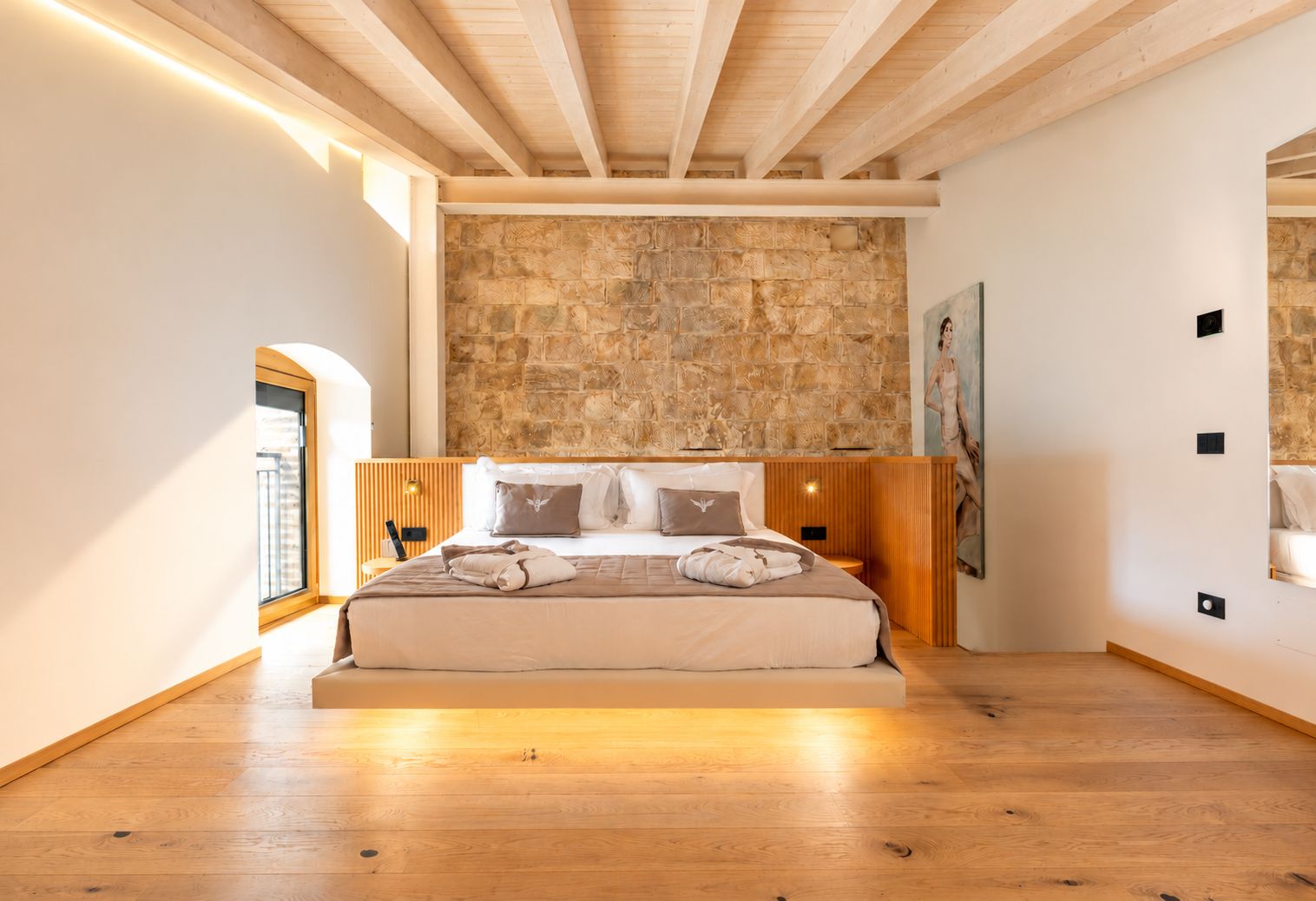 A bedroom with a stone wall, wooden ceiling beams, a bed with pillows, and a painting on the wall.