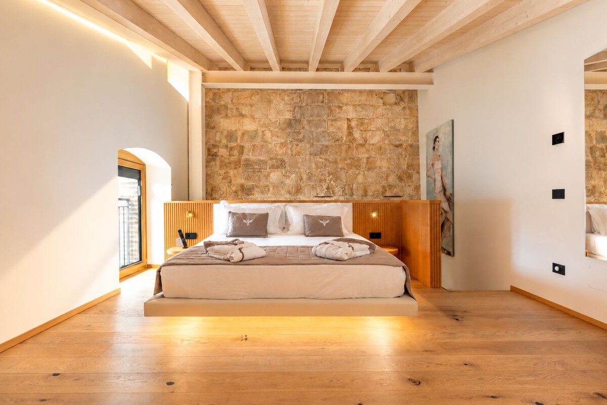 A bedroom with a stone wall, wooden ceiling beams, a bed with pillows, and a painting on the wall.