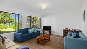 A spacious living room with a blue sofa, a wooden coffee table, and a TV on a stand. Large windows offer a view of the outdoors. - Rockpools in Mollymook (Mollymook Beach)