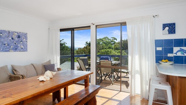 A spacious balcony with a wooden table and chairs, a view of greenery, and a kitchen area with blue tiles and a dolphin mural. - Rockpools in Mollymook (Mollymook Beach)