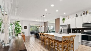 Una cucina moderna con mobili bianchi, una grande isola con sgabelli da bar, elettrodomestici in acciaio inossidabile e illuminazione a sospensione.