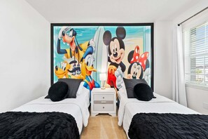 Habitación de hotel con dos camas, una mesita de noche y un mural grande con temática de Disney en la pared.