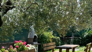Een houten picknicktafel en bankjes in een tuin met een stenen bouwwerk en bloeiende planten.