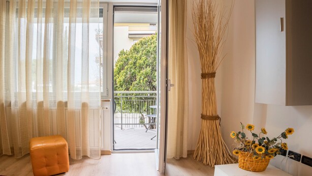 Een kamer met een schuifglasdeur naar een balkon, een rieten stoel, een klein oranje voetenbankje en een mand met zonnebloemen op een witte tafel.