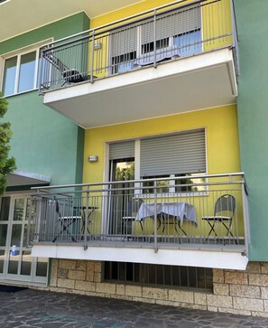 Un bâtiment à deux étages doté d’un balcon, d’une fenêtre avec un volet roulant et d’une petite table d’extérieur avec des chaises.