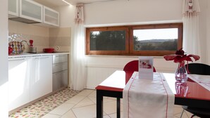 Una cocina con armarios blancos, una mesa de comedor con una silla roja y un ventanal con vistas al campo.