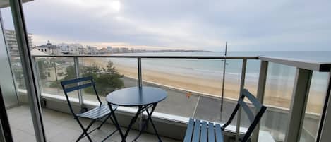 A balcony with two chairs and a table, overlooking a beach and cityscape.