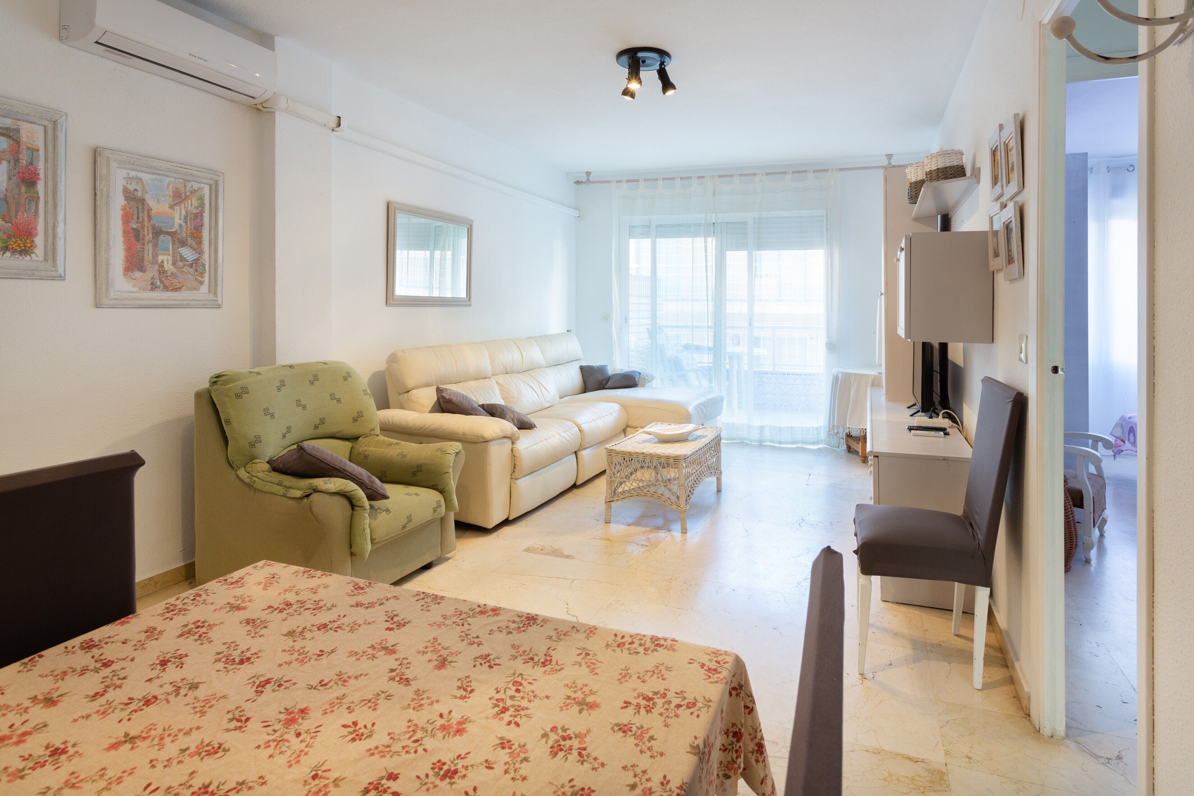 Ein helles Zimmer mit einer Couch, einem Sessel, einem kleinen Tisch und einem Bett mit blumigem Überwurf.