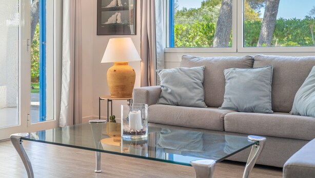 Een moderne woonkamer met een glazen salontafel, een beige bank met kussens, een lamp en uitzicht op bomen door grote ramen.
