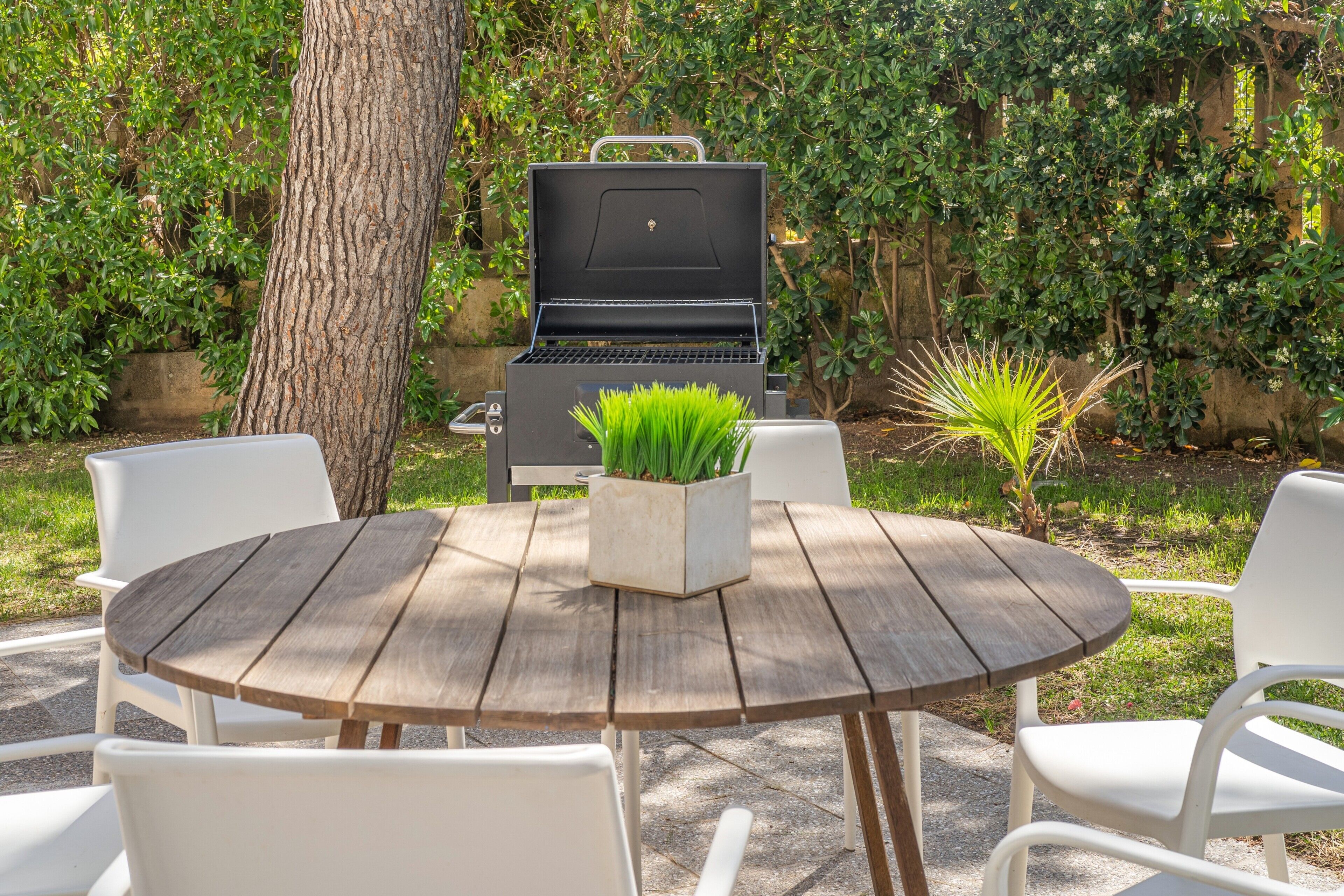 A round wooden table with white chairs, a barbecue grill, and a potted plant in the center.