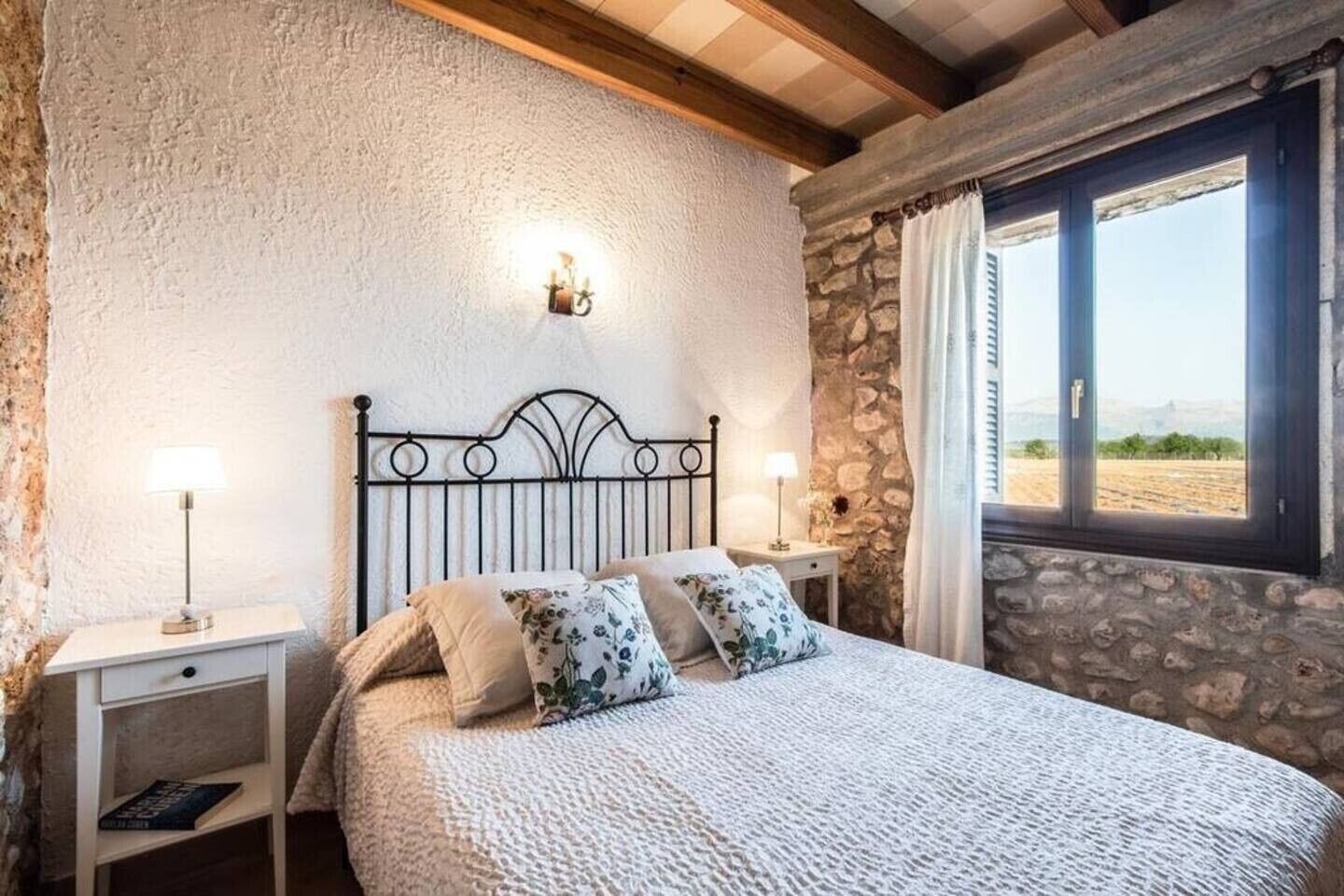 Un dormitorio con pared de piedra, cama con almohadas de flores, mesita de noche con lámpara y ventana con vistas.