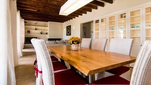 A dining area with a wooden table and wicker chairs, a hanging light fixture, and a kitchen with white cabinets in the background.