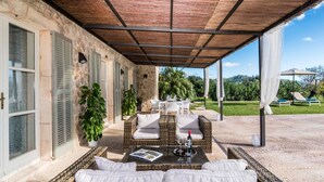 A covered outdoor seating area with wicker furniture, a table with glasses and a bottle, and a view of a lush garden and hills.