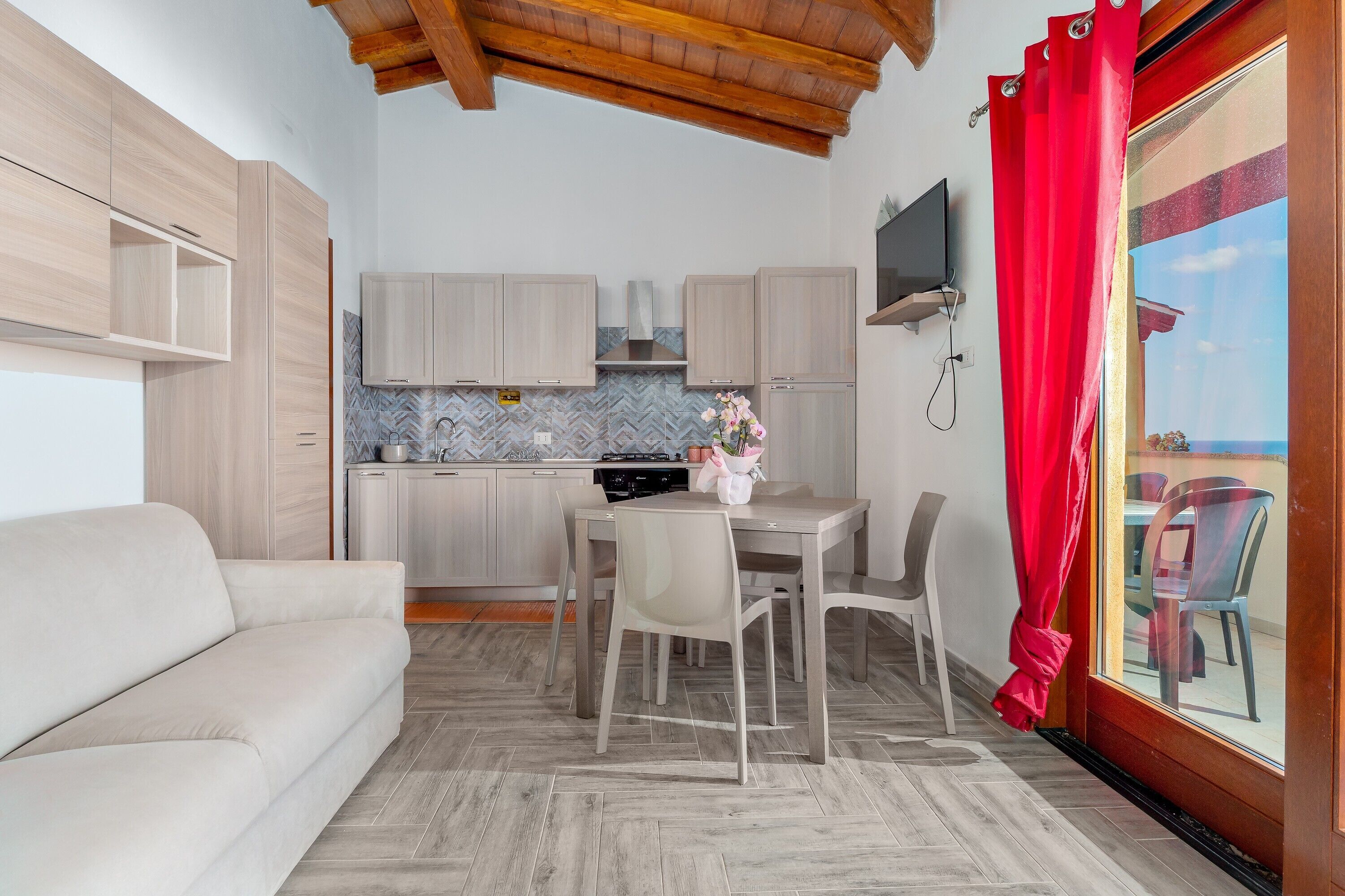 Cocina compacta con armarios de madera, una mesa de comedor con sillas, un sofá y un televisor montado en la pared.