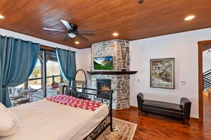 A bedroom with a bed, a stone fireplace, a television, and a view of the outdoors.