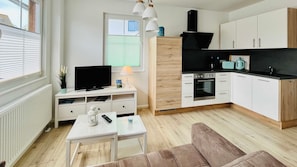 Une cuisine moderne avec des meubles blancs, une cuisinière noire et un plan de travail. On y trouve un téléviseur posé sur un meuble blanc, une petite table avec une lampe et une fenêtre avec des stores.