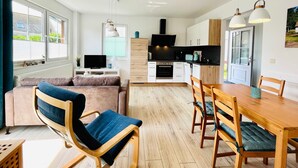 Ein modernes Wohnzimmer mit einer Couch, einem Holzes Esstisch mit Stühlen, einer Küchenzeile inklusive Herd und Backofen sowie einem Fernseher.