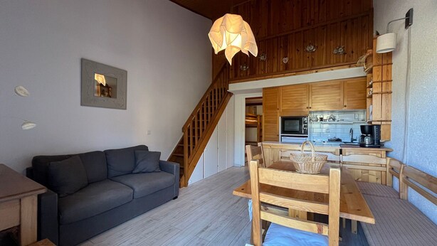 A cozy living room with a sofa, wooden dining table, and chairs. A staircase leads to a kitchen with wooden cabinets and a microwave. - Duplex T2 apartment on slope for 6 people with ski box in Super-Sauze (Enchastrayes)
