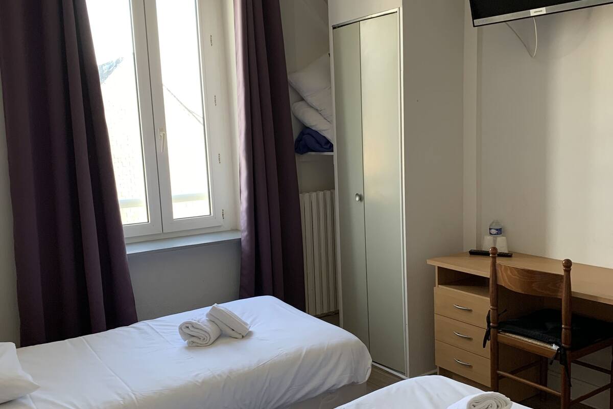 A hotel room with two single beds, a wooden desk, a television mounted on the wall, and a window with curtains.