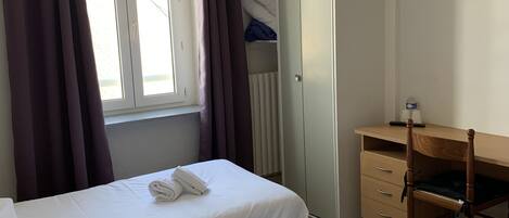 A hotel room with two single beds, a wooden desk, a television mounted on the wall, and a window with curtains.