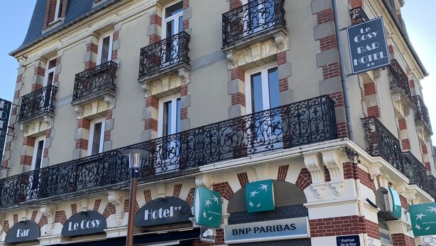 A hotel with a classic facade, featuring balconies with wrought iron railings, a ground-floor bar, and a visible ATM machine. - Le Cosy Riva Bella (Ouistreham)