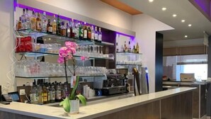 A modern bar area with a counter, shelves of bottles, glasses, and a pink orchid decoration. There are wooden stools and a seating area with a dark-colored bench. - Le Cosy Riva Bella (Ouistreham)