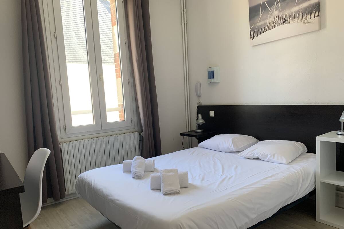 A neatly made bed with white linens and towels, a bedside table with a lamp, a chair, and a radiator.