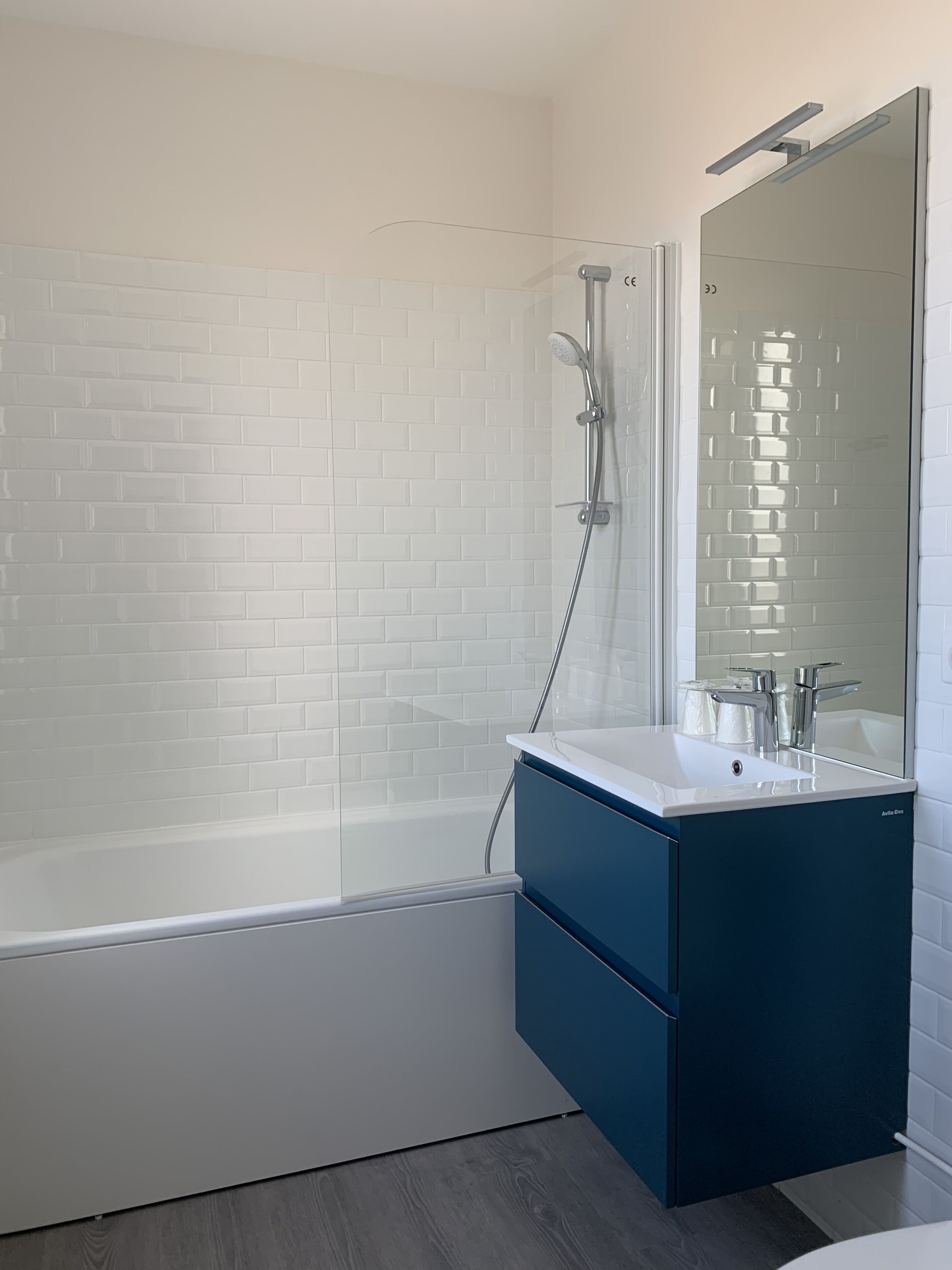 Une salle de bain moderne avec un mur de douche recouvert de carreaux blancs, une douche à l’italienne avec des parois en verre, un lavabo blanc et un meuble bleu, ainsi qu’un grand miroir.