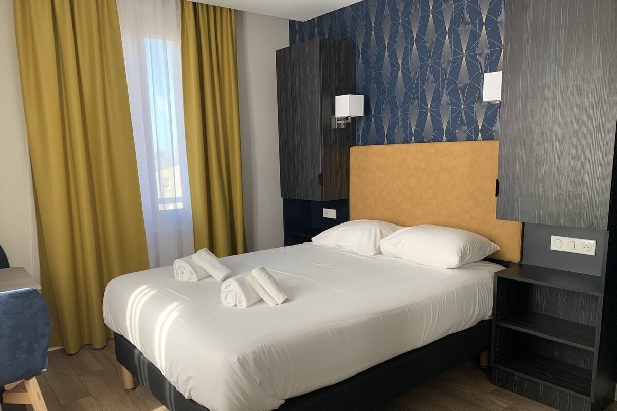 A neatly made bed with white linens and pillows, a wooden headboard, a bedside table with a lamp, and a wall with a geometric pattern.