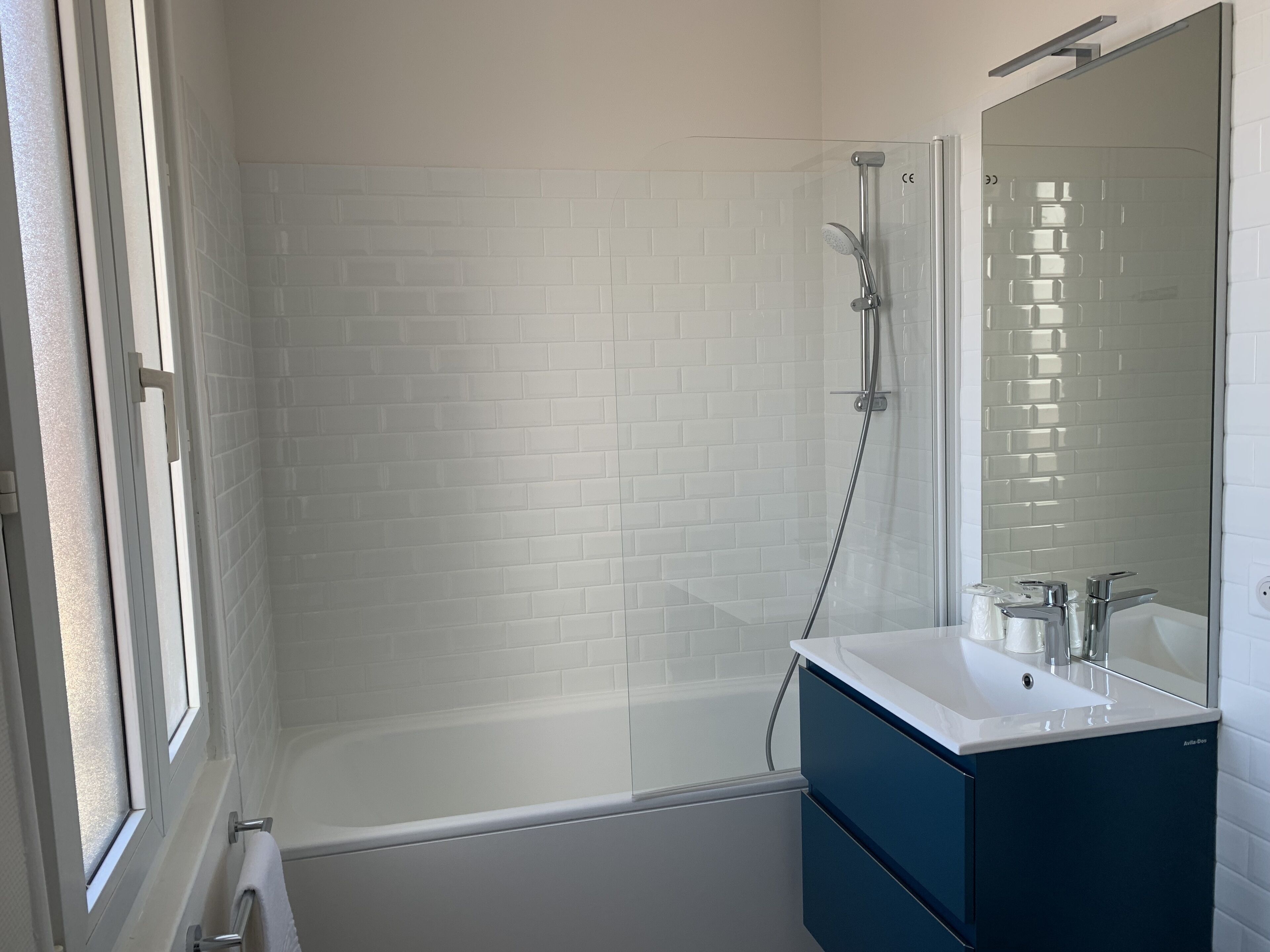 Un baño con ducha de azulejos blancos, bañera, lavabo con espejo y ventana.