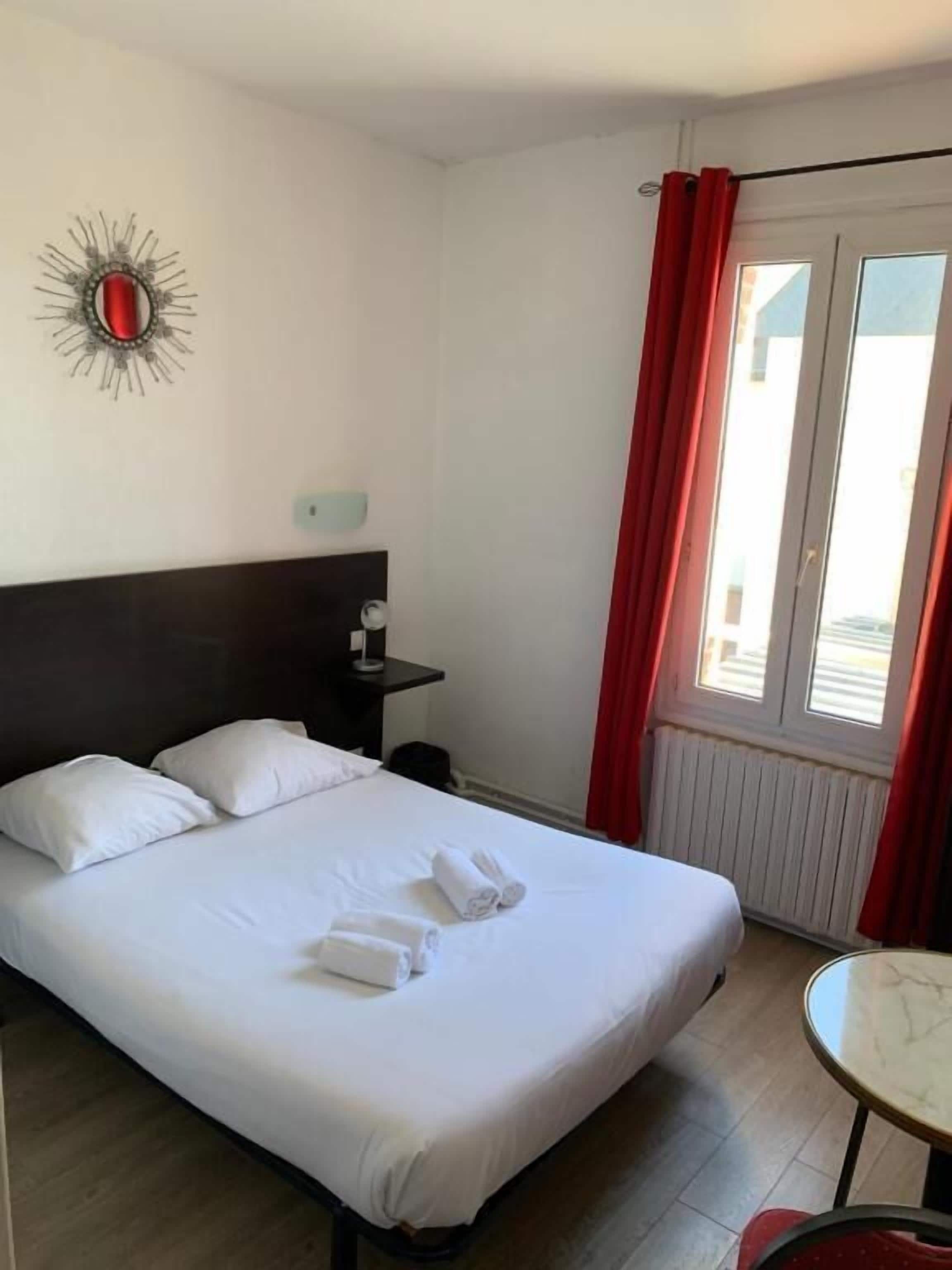 Une chambre d’hôtel avec un lit, des oreillers, une table de chevet, un élément de décoration murale, une fenêtre avec des rideaux rouges et une petite table.